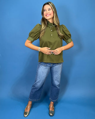 A woman modeling an olive green puff-sleeve blouse with a polka-dot collar and cuffs, paired with blue jeans and metallic shoes, standing against a solid blue studio backdrop.