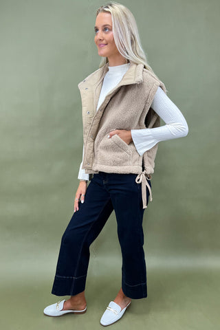 Woman wearing a beige vest over a white shirt with dark pants on a green background