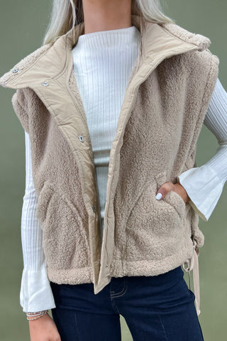 Beige fleece vest worn over a white sweater and dark jeans on a green background
