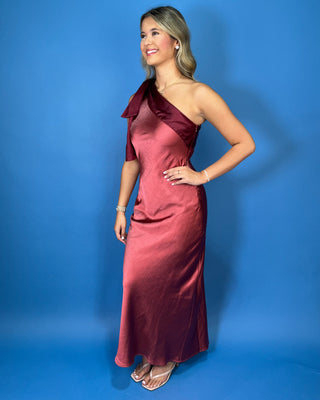 Burgundy satin one-shoulder maxi dress with statement bow detail, fitted silhouette, and elegant draped finish.