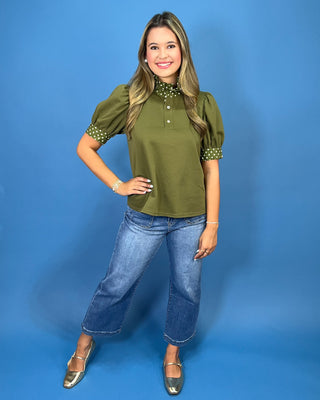 A woman modeling an olive green short-sleeve blouse with a high ruffled collar and matching polka-dot trim on the sleeves, paired with blue jeans and metallic shoes, against a solid blue studio background.