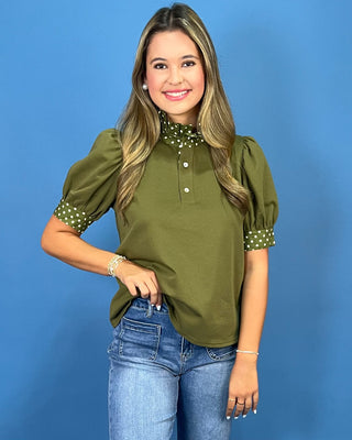 A woman modeling an olive green short-sleeve blouse with a polka-dot mock neck and cuffs, paired with blue jeans, against a solid blue studio background.