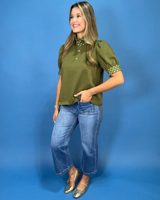A woman modeling an olive green puff-sleeve blouse with green and white polka-dot trim at the collar and cuffs, paired with blue jeans and metallic flats, against a solid blue studio background.