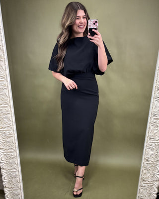 Woman taking a mirror selfie wearing a black outfit against an olive green background