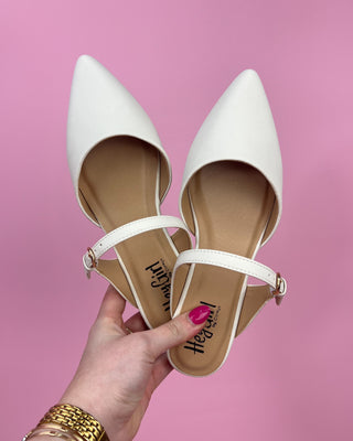 White pointed-toe shoes with ankle strap held by a hand against a pink background