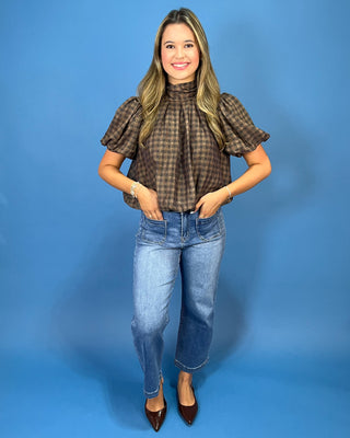 A woman modeling a brown and black checkered puff-sleeve mock neck blouse, paired with light-wash blue jeans and dark brown pointed-toe heels, standing against a solid blue studio backdrop.
