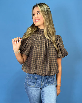 A woman with blonde-highlighted hair wearing a brown and black plaid short-sleeve blouse with puff sleeves and blue jeans, standing against a solid blue background.