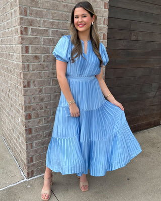 Woman wearing a blue pleated dress standing against a brick wall.