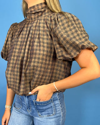 A woman wearing a brown and beige plaid puff-sleeve blouse tucked into blue jeans, standing against a solid blue background.