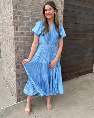 Woman in a blue dress standing against a brick wall.