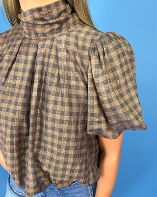 A close-up of a person wearing a brown and beige plaid mock neck top with puff sleeves, paired with blue jeans, set against a solid blue background.