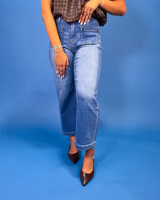 A person wearing medium-wash blue cropped jeans with rolled hems, styled with a brown plaid top and dark brown pointed-toe heels against a solid blue studio backdrop.