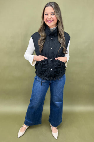 Woman wearing a black quilted vest over a white shirt with blue jeans on a green background