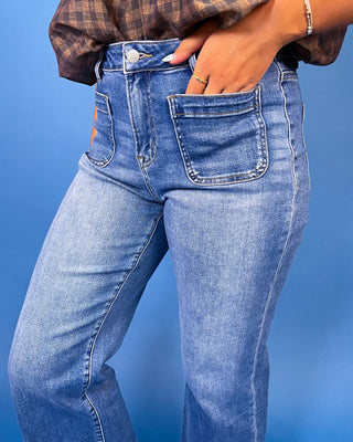 A close-up of a person wearing light blue denim jeans with a brown plaid shirt tucked into the waistband, standing against a solid blue background.