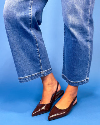 A person wearing blue cropped jeans and dark brown pointed slingback flats against a solid blue studio background.