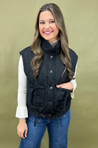 Woman wearing a black quilted vest over a white shirt with a green background