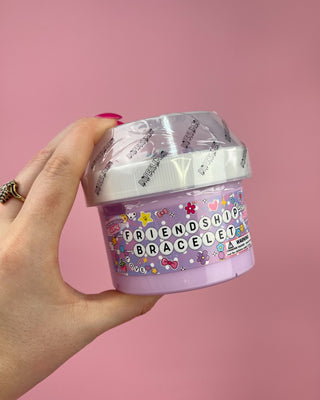Hand holding a container labeled 'Friendship Bracelet slime against a pink background