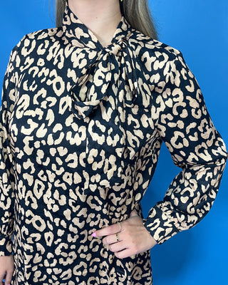 A woman wearing a long-sleeve leopard print blouse with a high neck and front tie detail, shown against a solid blue background.