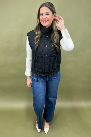 Woman wearing a black puffer vest over a white shirt and blue jeans on a green background