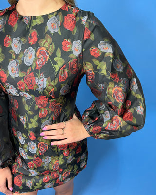A close-up of a person wearing a sheer black long-sleeve blouse with a red, gray, and green rose floral print
