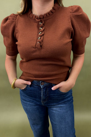 Person wearing a brown short-sleeve top with ruffled collar and blue jeans against a green background