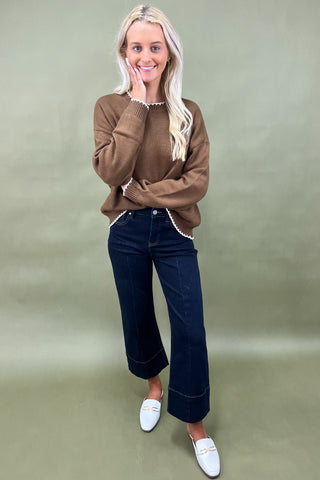 Woman wearing a brown sweater, dark jeans, and white loafers on a green background