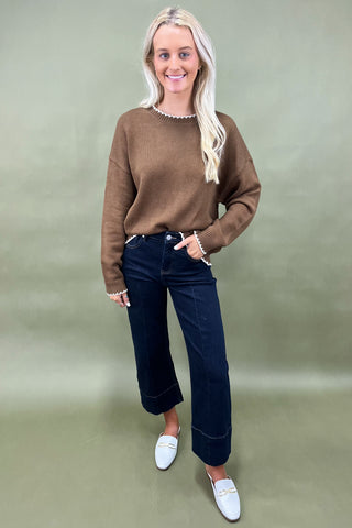 Woman wearing a brown sweater, dark jeans, and white loafers on a green background