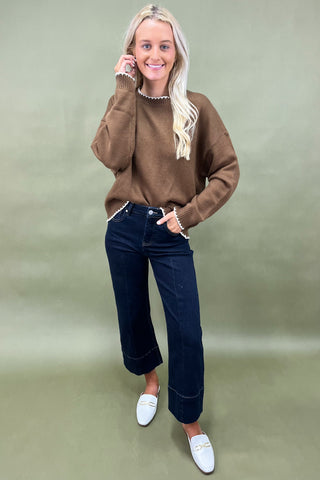 Woman wearing a brown sweater, dark jeans, and white loafers on a green background