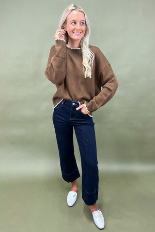 Woman wearing a brown sweater, dark jeans, and white loafers on a green background