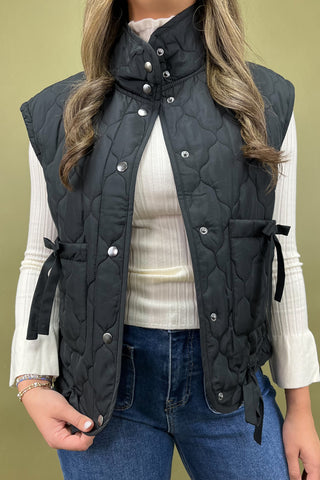 Person wearing a black quilted vest over a white shirt and blue jeans on a beige background