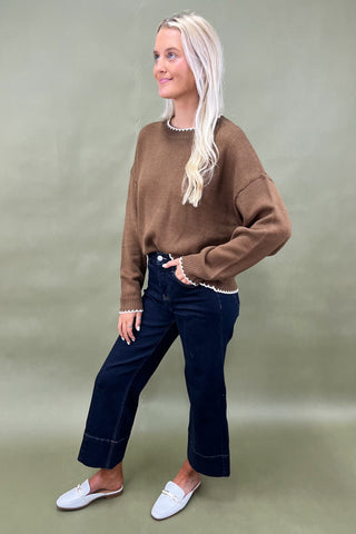 Woman wearing a brown sweater, dark jeans, and white loafers on a green background