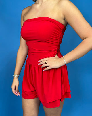 A woman wearing a bright red strapless ruched romper with short legs, standing against a solid blue background.