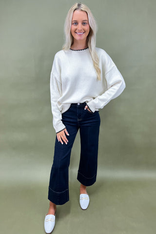 Woman wearing a white sweater and dark jeans against a green background