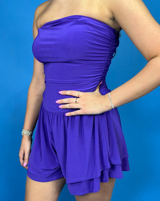 A woman wearing a strapless ruched romper in a vivid purple color, standing against a solid blue background.