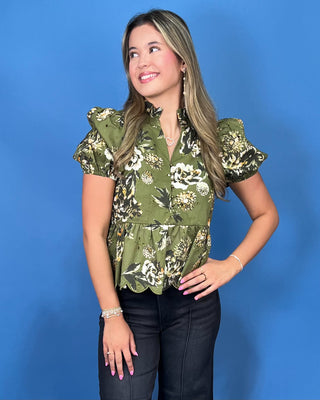 A woman modeling an olive green short-sleeve blouse with a white and black floral print, paired with black jeans