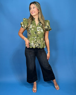 A woman modeling a green floral puff-sleeve blouse with a ruffled hem, paired with black wide-leg cropped pants and clear high-heel sandals
