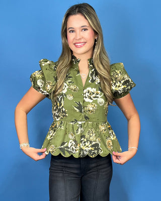 A woman modeling a short-sleeve green floral blouse with puff sleeves, a scalloped peplum hem, and a button-front neckline, paired with dark jean
