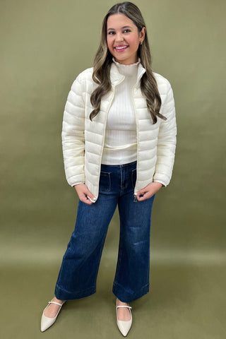 Woman wearing a white puffer jacket, white turtleneck, blue jeans, and white shoes on a green background