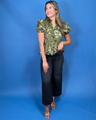 A woman modeling a short-sleeve olive green blouse with a white and black floral pattern, paired with black cropped jeans and metallic sandal