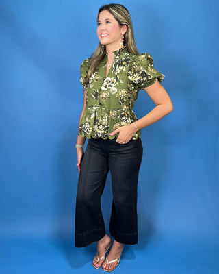 A woman modeling a short-sleeve peplum blouse with a green, black, and white floral pattern, paired with black cropped pants and white heeled sandal