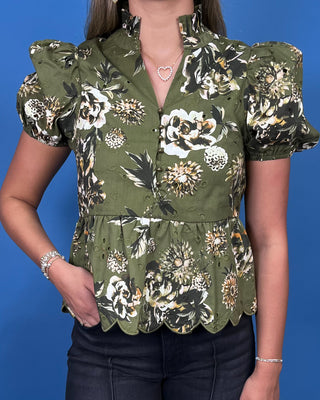 A woman wearing an olive green short-sleeve blouse with a bold floral print in white, beige, and black, featuring puffed sleeves and a scalloped peplum hem