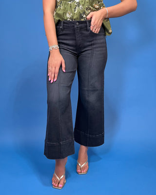A model wearing a pair of dark charcoal gray cropped wide-leg denim jeans with rolled hems, paired with a green floral top and metallic sandal