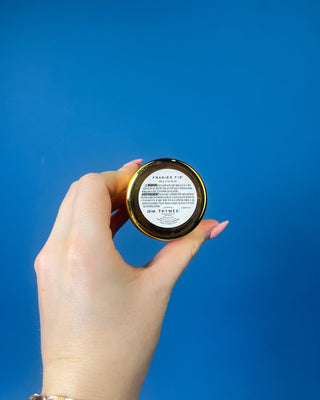 Thymes Frasier Fir Gold Votive candle displayed against a solid blue background. 