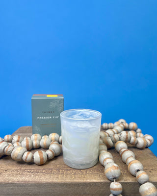 Frasier Fir Tidings Votive Candle in frosted marbled glass against a solid blue background. 