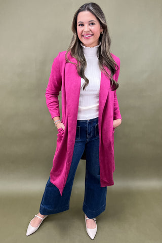 Woman wearing a pink coat, white shirt, blue jeans, and white shoes on a beige background