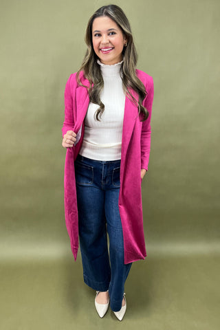 Woman wearing a pink coat, white sweater, blue jeans, and white shoes on a green background