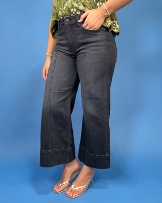 A person wearing dark charcoal gray wide-leg cropped jeans with rolled cuffs, paired with a green floral top and metallic flip-flop heel
