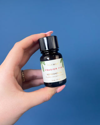 Thymes Frasier Fir Refresher Oil in amber glass bottle with signature evergreen label displayed on a solid blue background.