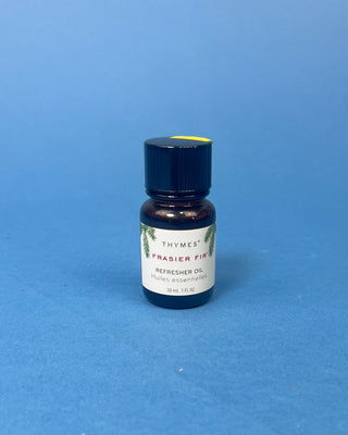 Thymes Frasier Fir Refresher Oil in amber glass bottle with signature evergreen label displayed on a solid blue background.