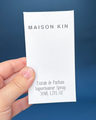 Hand holding a Maison Kin perfume packaging against a blue background